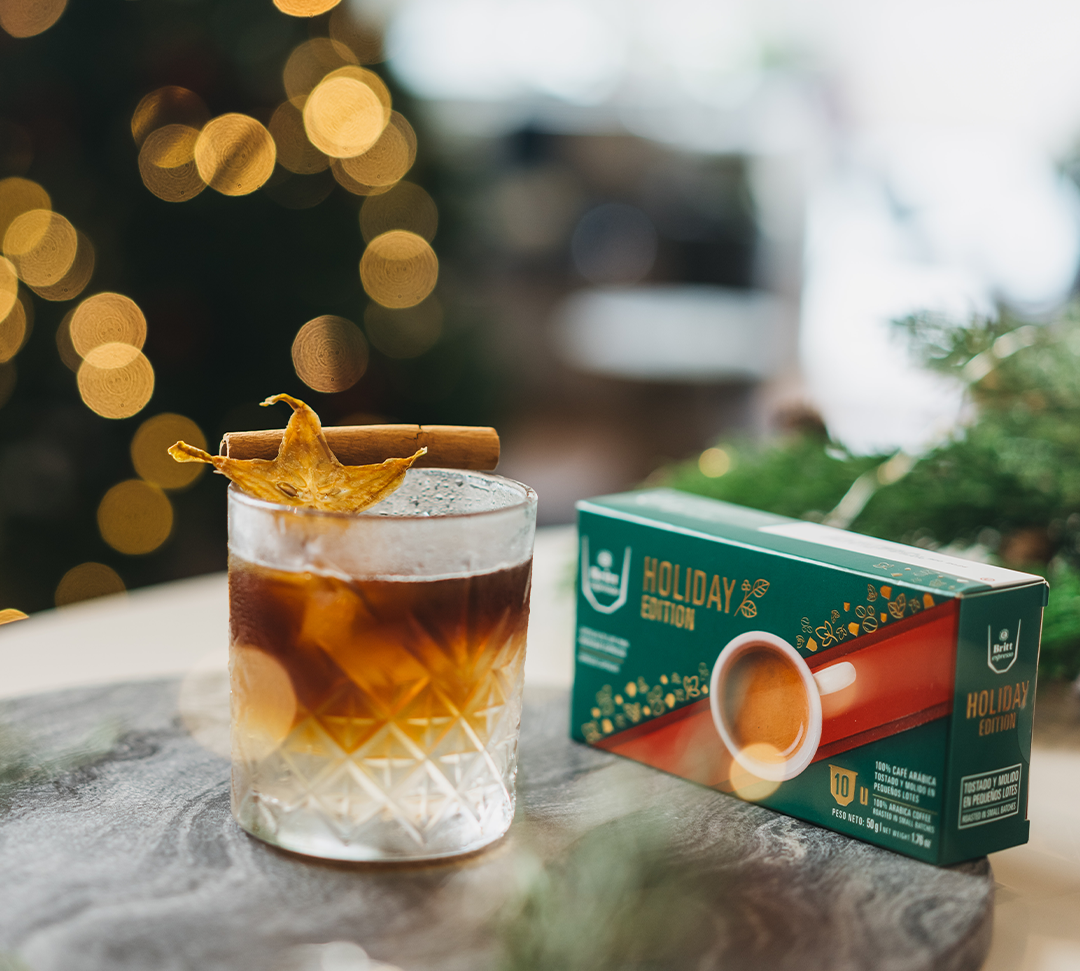 Ginger coffee tonic servido en vaso de vidrio y adornado con una astilla de canela y carambola deshidratada y una caja de cápsulas de Britt Espresso Holiday Edition 2025
