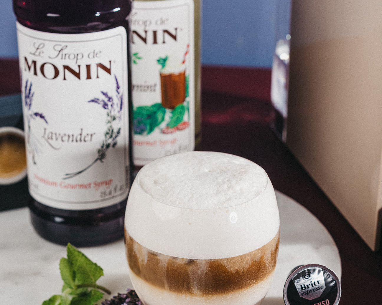 Vaso de Iced Lavender Mint Macchiato con espuma, sirope de lavanda Monin, menta fresca y cápsula Café Britt junto a una máquina de café.