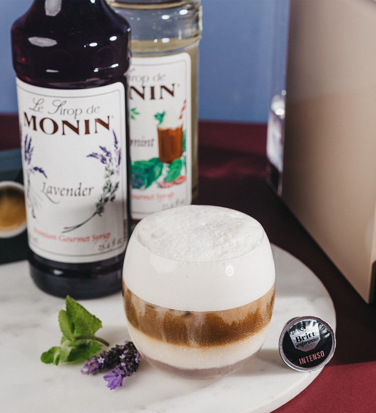 Vaso de Iced Lavender Mint Macchiato con espuma, sirope de lavanda Monin, menta fresca y cápsula Café Britt junto a una máquina de café.