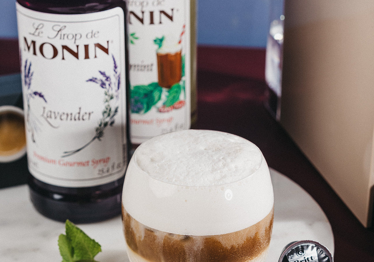 Vaso de Iced Lavender Mint Macchiato con espuma, sirope de lavanda Monin, menta fresca y cápsula Café Britt junto a una máquina de café.
