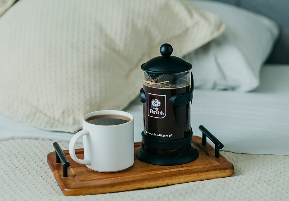 Una taza de café junto a una prensa francesa de Café Britt en una tabla de madera en una cama